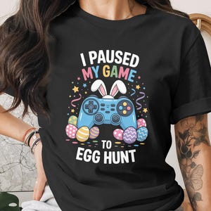Könnte beinhalten: Schwarzes T-Shirt mit einer bunten Grafik, die einen blauen Gamecontroller mit Hasenohren, Ostereiern und dem Text "I PAUSED MY GAME TO EGG HUNT" zeigt. Das Design umfasst Sterne und Konfetti.