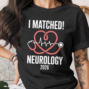 May include: Black t-shirt with the text "I MATCHED!" above a pink heart-shaped design incorporating a stethoscope and a heartbeat line. Below, it reads "NEUROLOGY 2026".