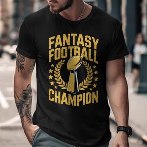 May include: Black t-shirt with a gold and white design. The shirt features the text "FANTASY FOOTBALL CHAMPION" with a trophy graphic, surrounded by a laurel wreath. The design has a distressed, vintage look.