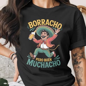 May include: Black t-shirt featuring a cartoon illustration of a man in a sombrero holding a beer bottle. The text "BORRACHO PERO BUEN MUCHACHO" is displayed in yellow and green. The man is wearing a red jacket and green pants.