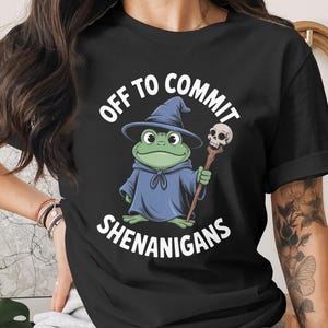 以下が含まれることがあります： 黒いTシャツ。漫画の魔法使い姿のカエルが、頭蓋骨の杖を持っています。「OFF TO COMMIT SHENANIGANS」の文字がプリントされています。カエルは青いローブと帽子を着用。面白くて気まぐれなデザインです。