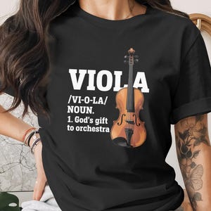 May include: Black t-shirt featuring the word "VIOLA" in white, with a definition and a drawing of a violin. The definition includes the pronunciation and the phrase "God's gift to orchestra."