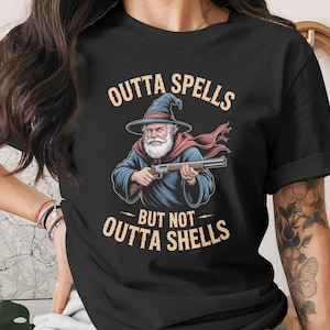 May include: Black t-shirt featuring a wizard illustration holding a rifle. The wizard wears a blue hat and robe with a red scarf. Text above reads "OUTTA SPELLS" and below "BUT NOT OUTTA SHELLS".