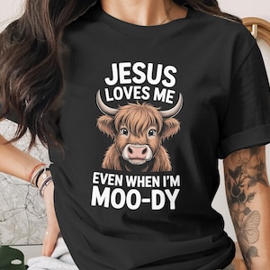 May include: Black t-shirt featuring a brown Highland cow illustration with the text "JESUS LOVES ME EVEN WHEN I'M MOO-DY" in white. The cow has large horns and a fluffy coat. The shirt is a casual wear item.