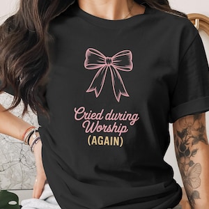 Puede incluir: Camiseta negra con un lazo rosa y el texto "Cried during Worship (AGAIN)". El texto está en cursiva con un acento amarillo. Una prenda informal.