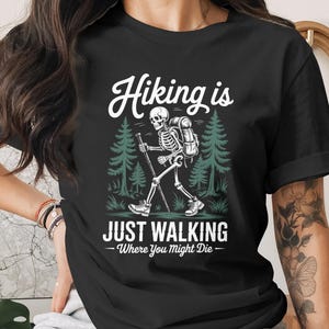 May include: Black t-shirt with a graphic of a skeleton hiking in a forest. The text reads "Hiking is Just Walking Where You Might Die". The design features a skeleton with a backpack and hiking stick.
