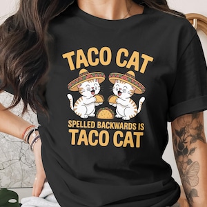 Puede incluir: Camiseta negra con un diseño de dibujos animados de dos gatos con sombreros, cada uno sosteniendo un taco. El texto dice "TACO CAT" y "SPELLED BACKWARDS IS TACO CAT" en amarillo. Un diseño divertido con temática de comida.