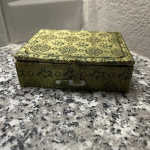 May include: A small, square, green and gold box with a floral pattern. The box has a clasp on the front.