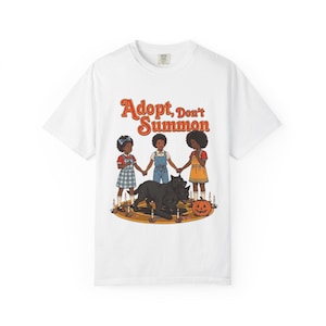 Puede incluir: Una camiseta blanca de manga corta con un gráfico de tres niños tomados de la mano alrededor de un perro negro de tres cabezas y una calabaza sobre una alfombra circular con velas. El texto Adopt, Don't Summon aparece en letras naranjas.