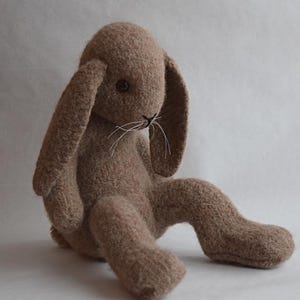 Op de afbeelding: Een bruin pluchen konijn met lange oren en een zittende houding. Het knuffeldier heeft een getextureerd, fuzzy uiterlijk, met een donker oog en witte snorharen. De poten van het konijn zijn uitgestrekt en het lijkt te rusten.