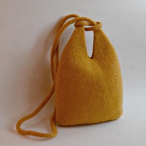 May include: Mustard-yellow felt bag with a knotted strap. The bag has a rounded, bell-like shape with a cutout at the top. The strap is made of the same felt material. The bag is set against a white background.