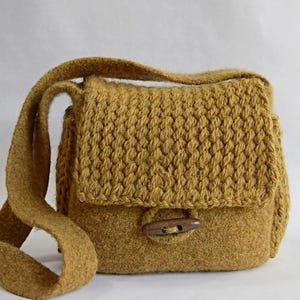 May include: Mustard-coloured, handmade shoulder bag with a textured, knitted flap and a wooden toggle closure. The bag features a long, matching strap for crossbody or shoulder wear. The bag is made of a soft, wool-like material.