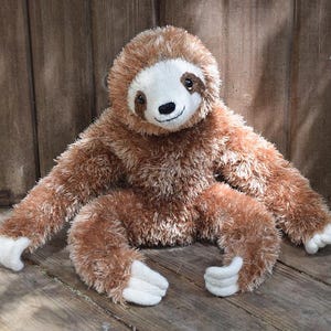 May include: A plush sloth toy with brown fur, a white face, and a small black nose. The sloth has long arms and legs with white paws. The toy is sitting on a wooden surface.