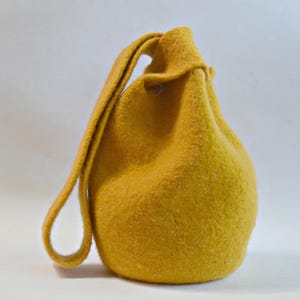 Könnte beinhalten: Senfgelbe Filz-Kordelzugtasche mit Schlaufengriff. Die Tasche hat eine abgerundete Form und eine strukturierte Oberfläche. Der Griff ist oben an der Tasche befestigt. Die Tasche ist aus weichem Filz gefertigt.