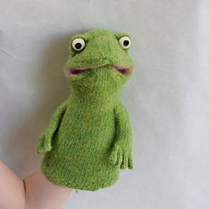 May include: A green knitted frog hand puppet with large, round eyes and a pink mouth. The puppet has small arms and is designed to fit over a hand. The frog puppet is made of a textured, green yarn.