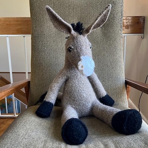 May include: A handmade plush donkey toy with a light brown body, long ears, and black accents on the mane, hooves, and tail. The donkey has a white muzzle and is sitting on a chair.