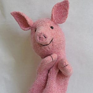 May include: A pink pig hand puppet with large ears, a smiling face, and small black eyes. The puppet's arms are extended, and the fingers have black stitching. The puppet is made of a textured, soft material.