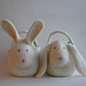 May include: Two white felt bunny bags with black eyes and pink noses. One bunny has upright ears, the other has floppy ears. Both have a small handle and a green interior. These bags are perfect for Easter.