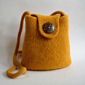 Może przedstawiać: Musztardowa filcowa torba na ramię z klapką i dużym, ciemnobrązowym guzikiem. Torba ma długi, pasujący pasek i zaokrąglony, cylindryczny kształt. Filc ma teksturowany wygląd.
