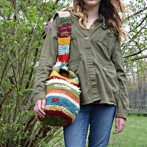 Könnte beinhalten: Eine bunte, handgefertigte Umhängetasche mit einem Streifenmuster in Rot-, Blau-, Gelb- und Grüntönen. Die Tasche hat einen dicken, strukturierten Riemen und eine abgerundete Form. Die Tasche besteht aus einem gestrickten oder gehäkelten Material.