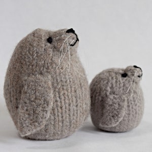 May include: Two knitted gray seal plushies, one larger than the other, with black eyes and white whiskers.