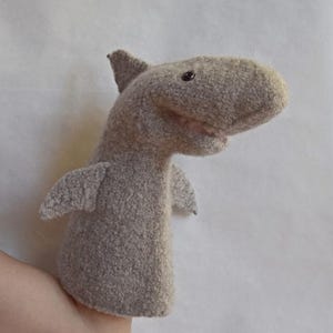 May include: A gray shark hand puppet made of soft fabric. The puppet has a fin on its back, small side fins, and a smiling mouth. The shark's eyes are small and dark. The puppet is designed to fit over a hand.