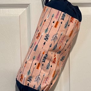 May include: A fabric storage bag with a drawstring closure, hanging from a door. The bag features a pattern of arrows in various colors, including pink, orange, and blue, against a light pink background. The top and bottom are navy blue.