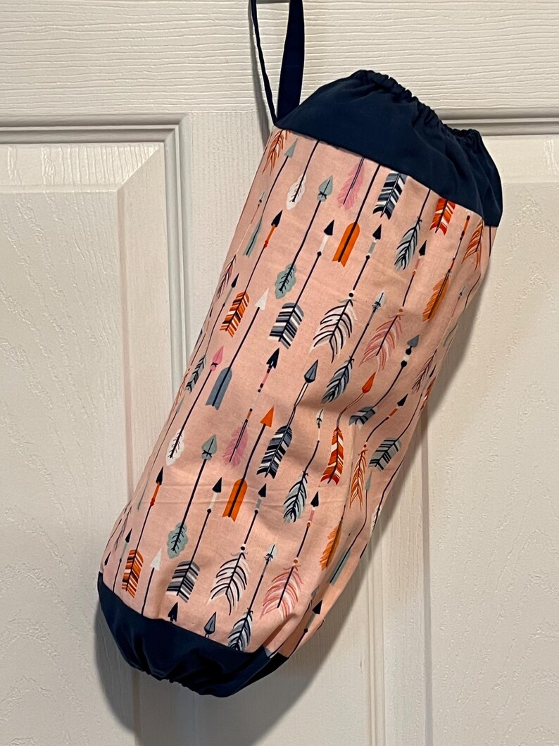 May include: A fabric storage bag with a drawstring closure, hanging from a door. The bag features a pattern of arrows in various colors, including pink, orange, and blue, against a light pink background. The top and bottom are navy blue.