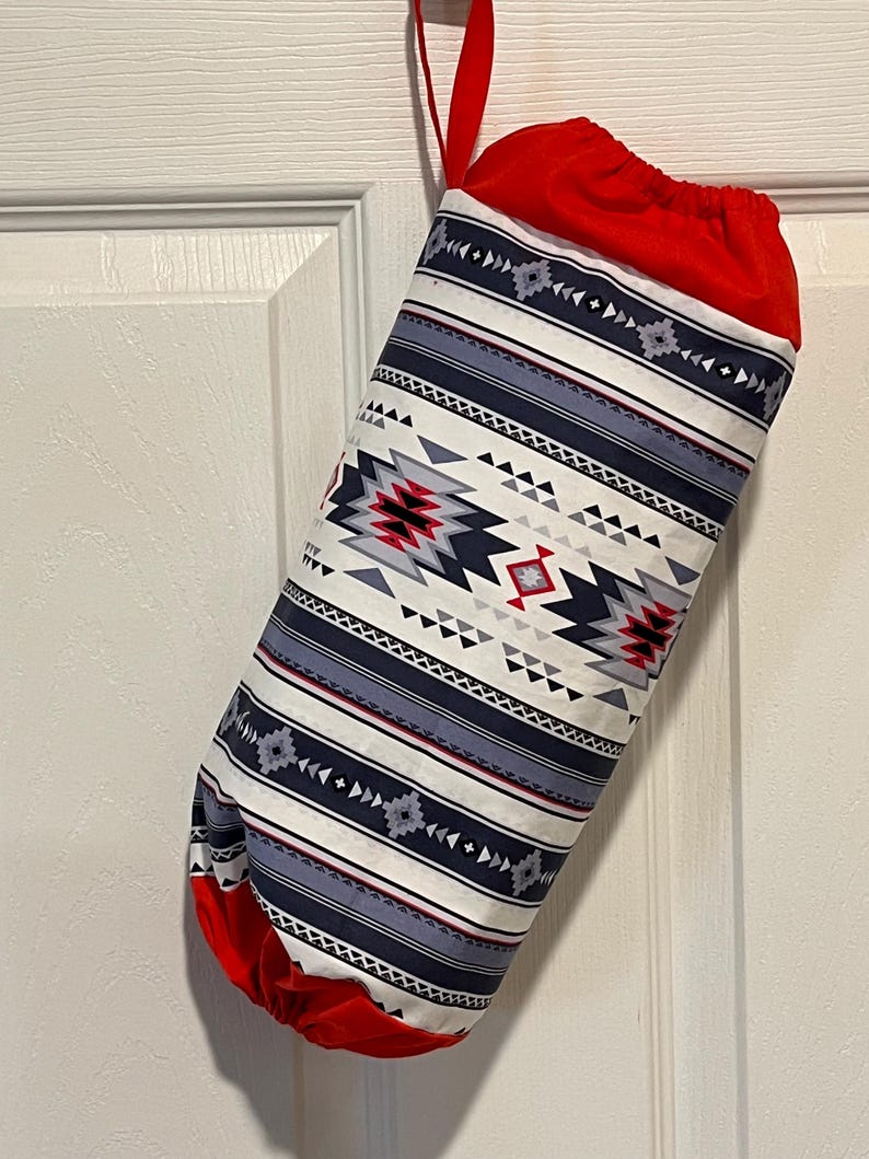 May include: A fabric stocking with a Southwestern pattern in white, navy, red, and gray. The top and bottom are red, and a red ribbon is attached for hanging. The stocking is hanging on a white door.