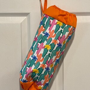 May include: A fabric bag with a colorful cactus print, featuring green, pink, and orange cacti with flowers on a white background. The top and bottom are solid orange, and it has a matching orange loop for hanging.