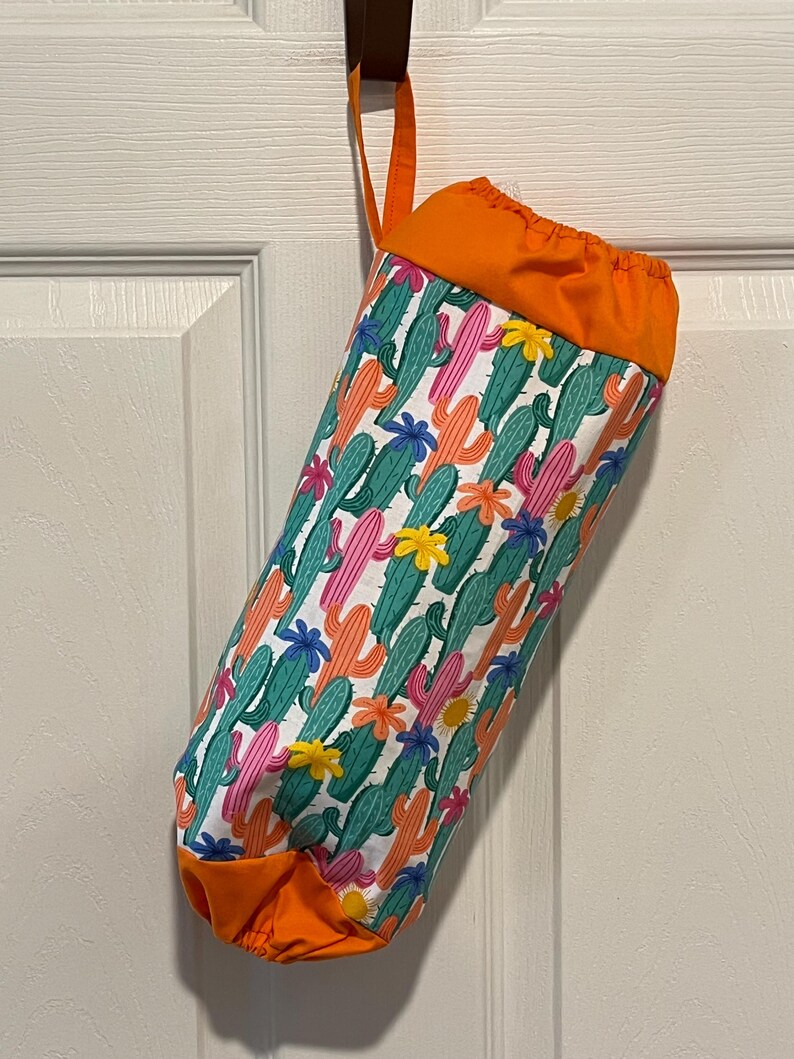 May include: A fabric bag with a colorful cactus print, featuring green, pink, and orange cacti with flowers on a white background. The top and bottom are solid orange, and it has a matching orange loop for hanging.