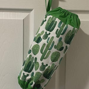 May include: A fabric bag with a green top and loop, hanging on a door. The bag is white with a repeating pattern of green cacti. The bottom of the bag is also green, and a plastic bag is visible inside.