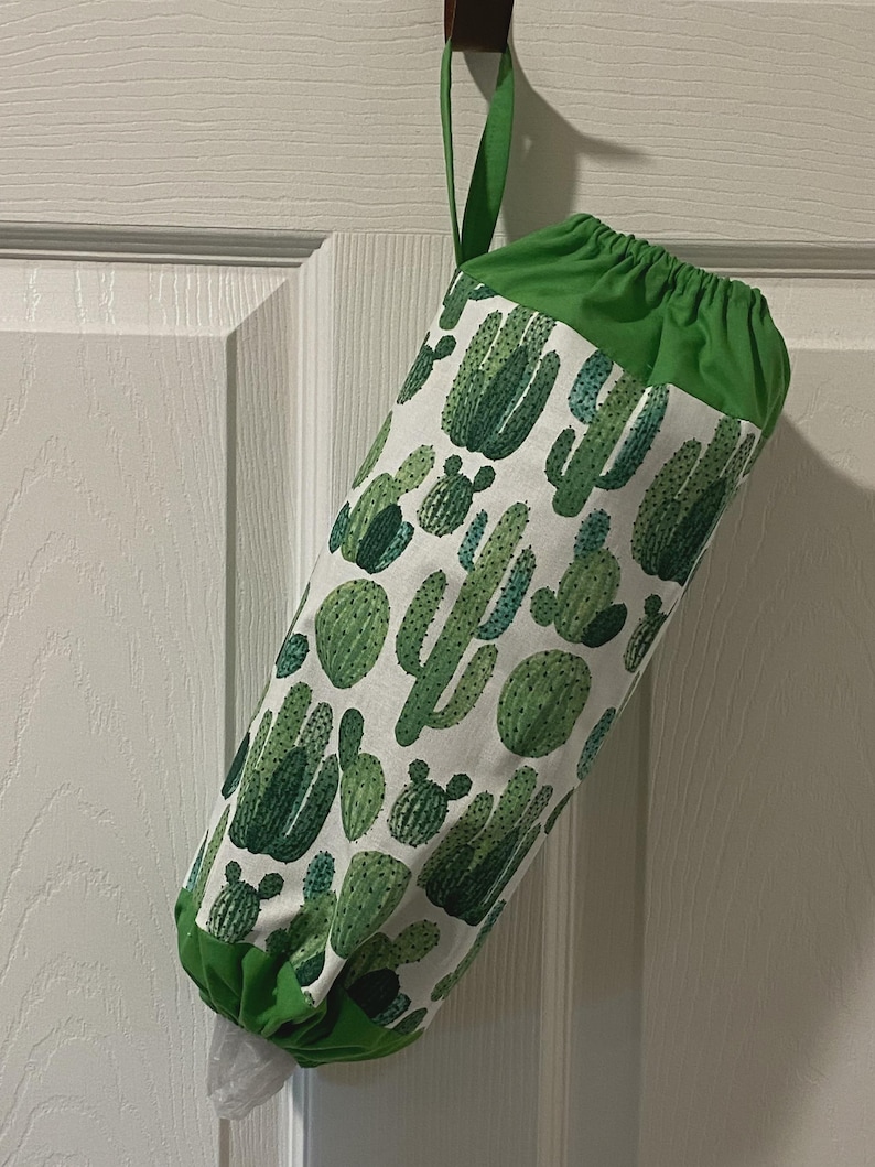 May include: A fabric bag with a green top and loop, hanging on a door. The bag is white with a repeating pattern of green cacti. The bottom of the bag is also green, and a plastic bag is visible inside.