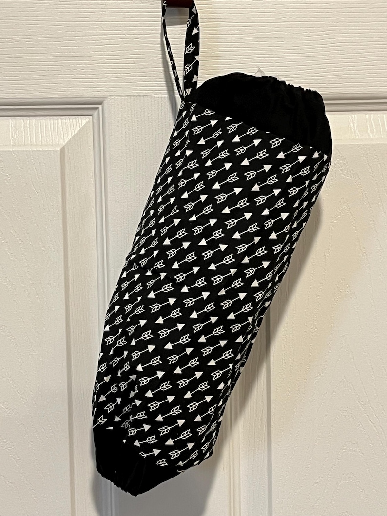 May include: A black and white fabric bag with a drawstring closure, hanging from a door. The bag is cylindrical and patterned with white arrows on a black background. The top and bottom are solid black.