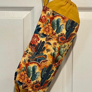 May include: A fabric bag with a yellow drawstring top and loop. The bag features a colorful pattern of cacti and flowers in shades of orange, blue, and yellow. The bag is hanging on a white door.