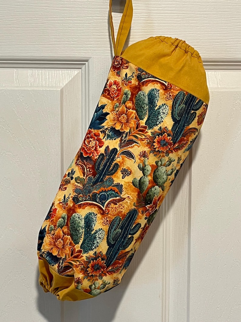 May include: A fabric bag with a yellow drawstring top and loop. The bag features a colorful pattern of cacti and flowers in shades of orange, blue, and yellow. The bag is hanging on a white door.