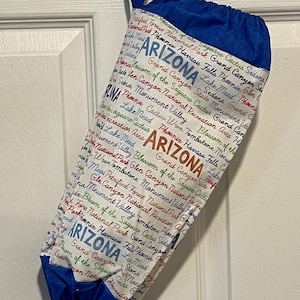 May include: A white and blue drawstring bag featuring the word "ARIZONA" in large letters, along with various Arizona landmarks and place names. The bag has a blue top and a blue loop for hanging.