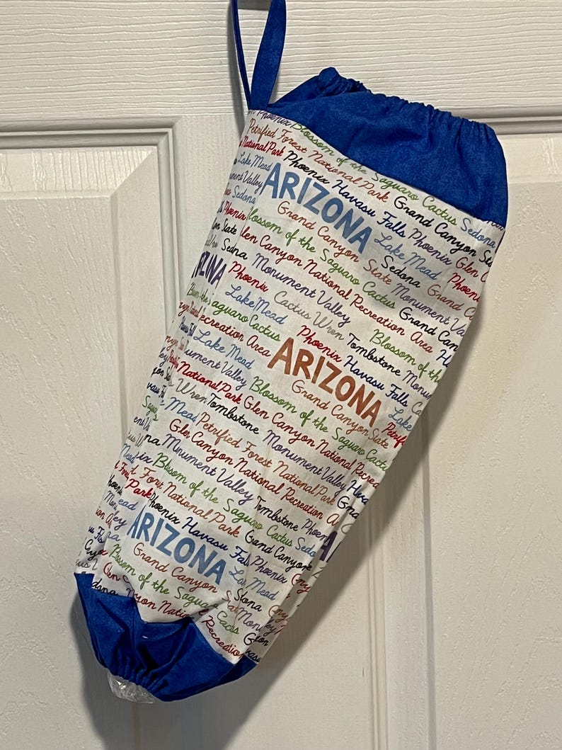 May include: A white and blue drawstring bag featuring the word "ARIZONA" in large letters, along with various Arizona landmarks and place names. The bag has a blue top and a blue loop for hanging.