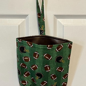 May include: A green fabric bag with a football and helmet pattern, hanging from a brown door hook. The bag features a repeating pattern of brown footballs and black helmets on a green background with white stripes.