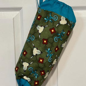 May include: A fabric bag with a teal top and bottom, and a teal hanging loop. The bag's body is olive green with a repeating pattern of white bull skulls, turquoise lizards, and red and yellow geometric shapes.