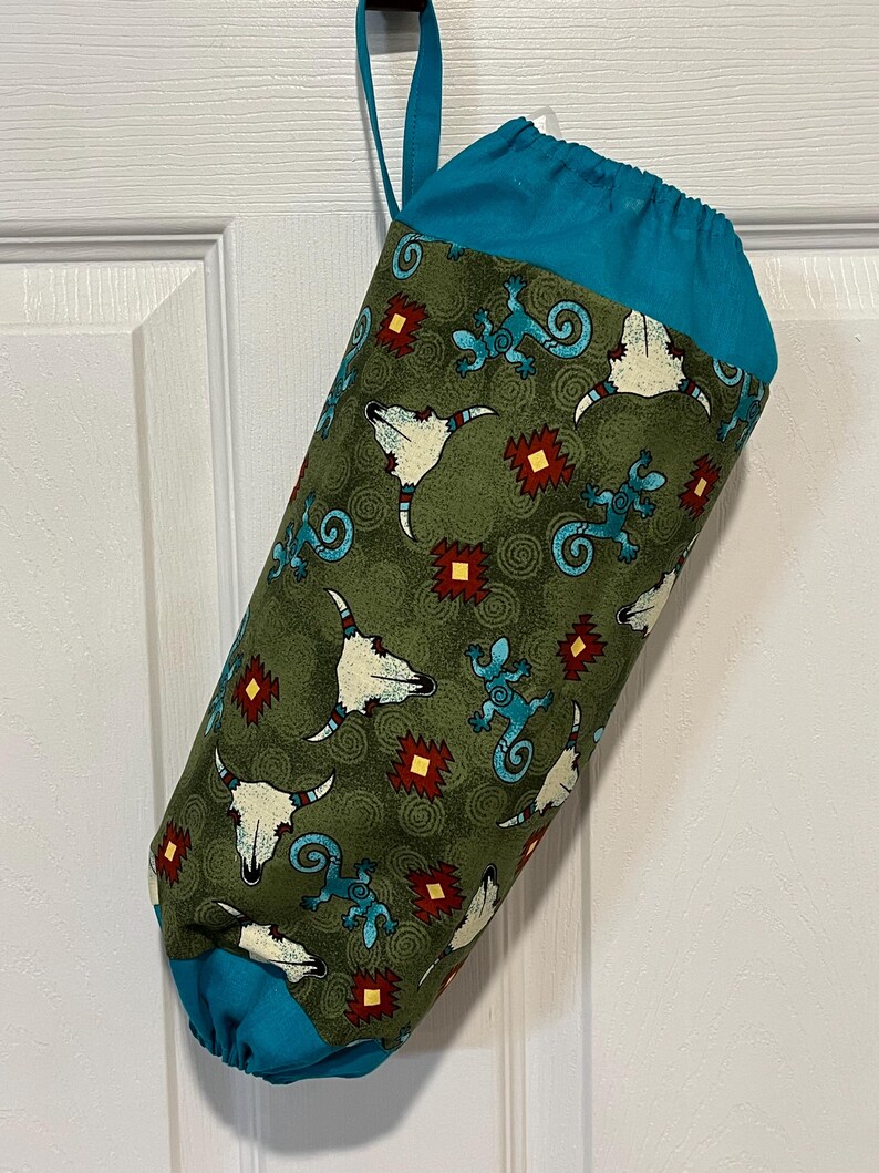 May include: A fabric bag with a teal top and bottom, and a teal hanging loop. The bag's body is olive green with a repeating pattern of white bull skulls, turquoise lizards, and red and yellow geometric shapes.