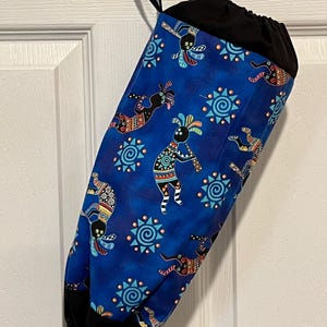 May include: A blue fabric bag with a black drawstring top and a black hanging loop. The bag is patterned with Kokopelli figures and sun symbols in various colors. The bag is hanging against a white door.