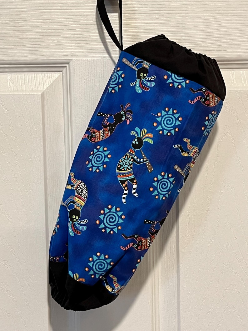 May include: A blue fabric bag with a black drawstring top and a black hanging loop. The bag is patterned with Kokopelli figures and sun symbols in various colors. The bag is hanging against a white door.