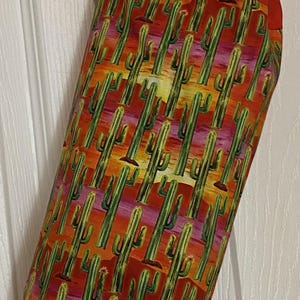 May include: A fabric bag with a vibrant cactus print in shades of green, orange, and yellow. The bag has a solid red top and a red loop for hanging. The bag is hanging on a white door.