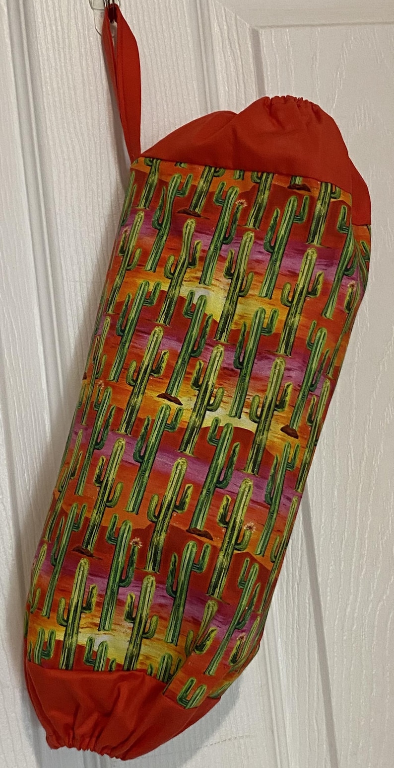 May include: A fabric bag with a vibrant cactus print in shades of green, orange, and yellow. The bag has a solid red top and a red loop for hanging. The bag is hanging on a white door.