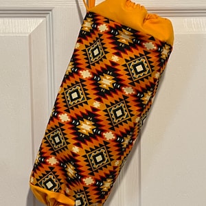May include: A fabric bag with a drawstring closure and a loop for hanging. The bag features a geometric pattern in shades of orange, black, and white. The top and bottom of the bag are solid orange.