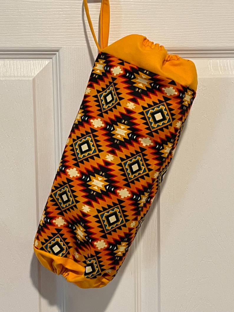 May include: A fabric bag with a drawstring closure and a loop for hanging. The bag features a geometric pattern in shades of orange, black, and white. The top and bottom of the bag are solid orange.