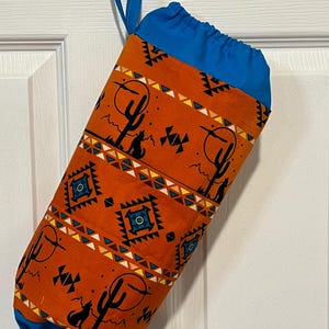 May include: Orange fabric bag with a blue drawstring top and handle. The bag features a Southwestern desert design with cacti, geometric patterns, and a howling wolf silhouette. The bag is hanging on a white door.