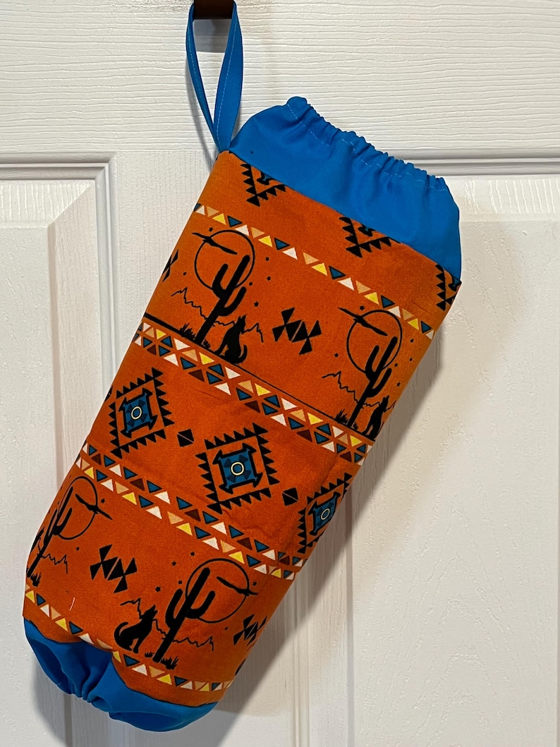 May include: Orange fabric bag with a blue drawstring top and handle. The bag features a Southwestern desert design with cacti, geometric patterns, and a howling wolf silhouette. The bag is hanging on a white door.