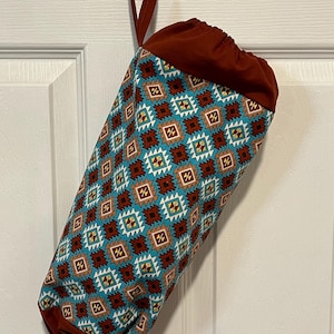 May include: A cylindrical fabric bag with a geometric pattern in teal, brown, and white. The bag has a drawstring closure and a loop for hanging. The bag is hanging on a brown hook.